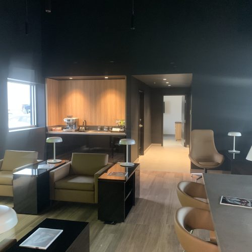 Poplar Painting - A modern lounge area with black walls, featuring a variety of seating options such as armchairs and bar stools, a wooden service counter with a coffee machine, and natural light entering through a window.